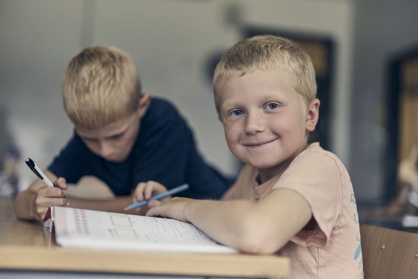 Två elever på Noblaskolan sitter bredvid varandra under en lektion, den ena tittar glatt in i kameran.