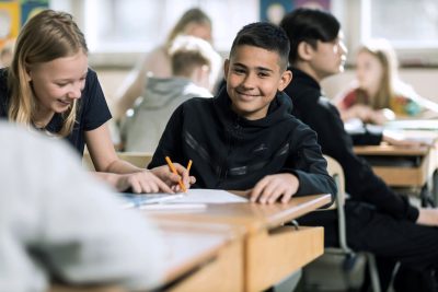 En elev på Noblaskolan sitter i ett klassrum, kollar in i kameran och ler.
