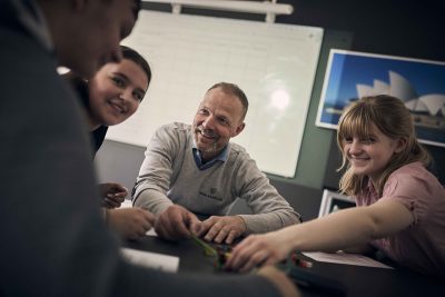 Tre elever på Noblaskolan sitter i klassrummet ihop med en lärare.