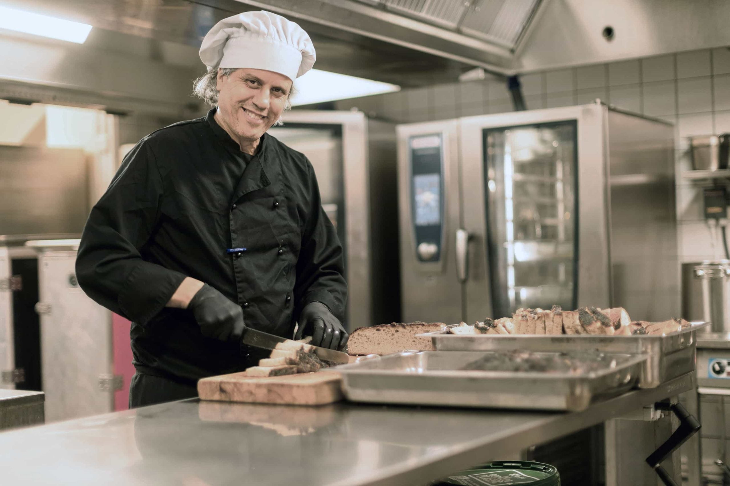 Noblaskolans kock står i köket och skär bröd, ler in i kameran.